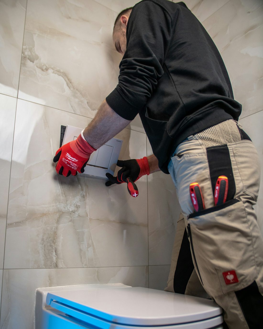 Plumber in work gear repairing a wall-mounted toilet cistern in a tiled bathroom