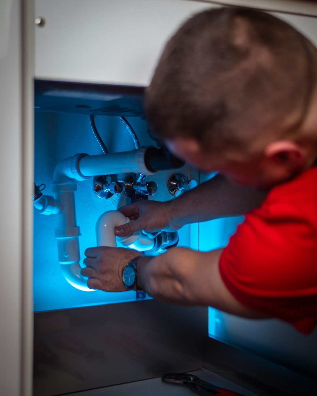 Plumber's hand turning a valve on pipework beneath a sink, task-lit in blue